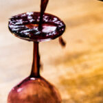 particolare del collo di una bottiglia di vino mentre ne viene versato fuori il contenuto sopra un calice appoggiato al contrario su un tavolo di legno