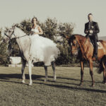 Due sposi fanno una passeggiata a cavallo all'interno di una tenuta. Quello di lei è biano come il suo vestito, mentre quello di lui è marrone.