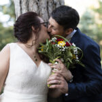 Piano medio di una coppia di sposi che si bacia davanti al tronco di un albero in un parco di una villa. Entrambi tengono con una mano il bouquet della sposa davanti a loro