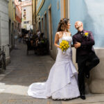Figura intera di una coppia di sposi in una via del centro della Città Vecchia a Trieste. Sono entrambe in piedi e si guardano negli occhi. Lei ha il bouquet in mano. Lui è appoggiato con la schiena e con un piede a u muro