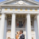 Piano medio di una coppia di sposi fotografata leggermente dal basso mentre si bacia tra le colonne del palazzo della Borsa a Trieste. Davanti a loro appoggiato sul bordo della fontana antistante, il bouquet della sposa