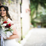 Piano medio di una sposa sorridente appoggiata a un muro di pietra all'interno del parco di una villa. in una mano tiene il bouquet di rose bianche e rosse