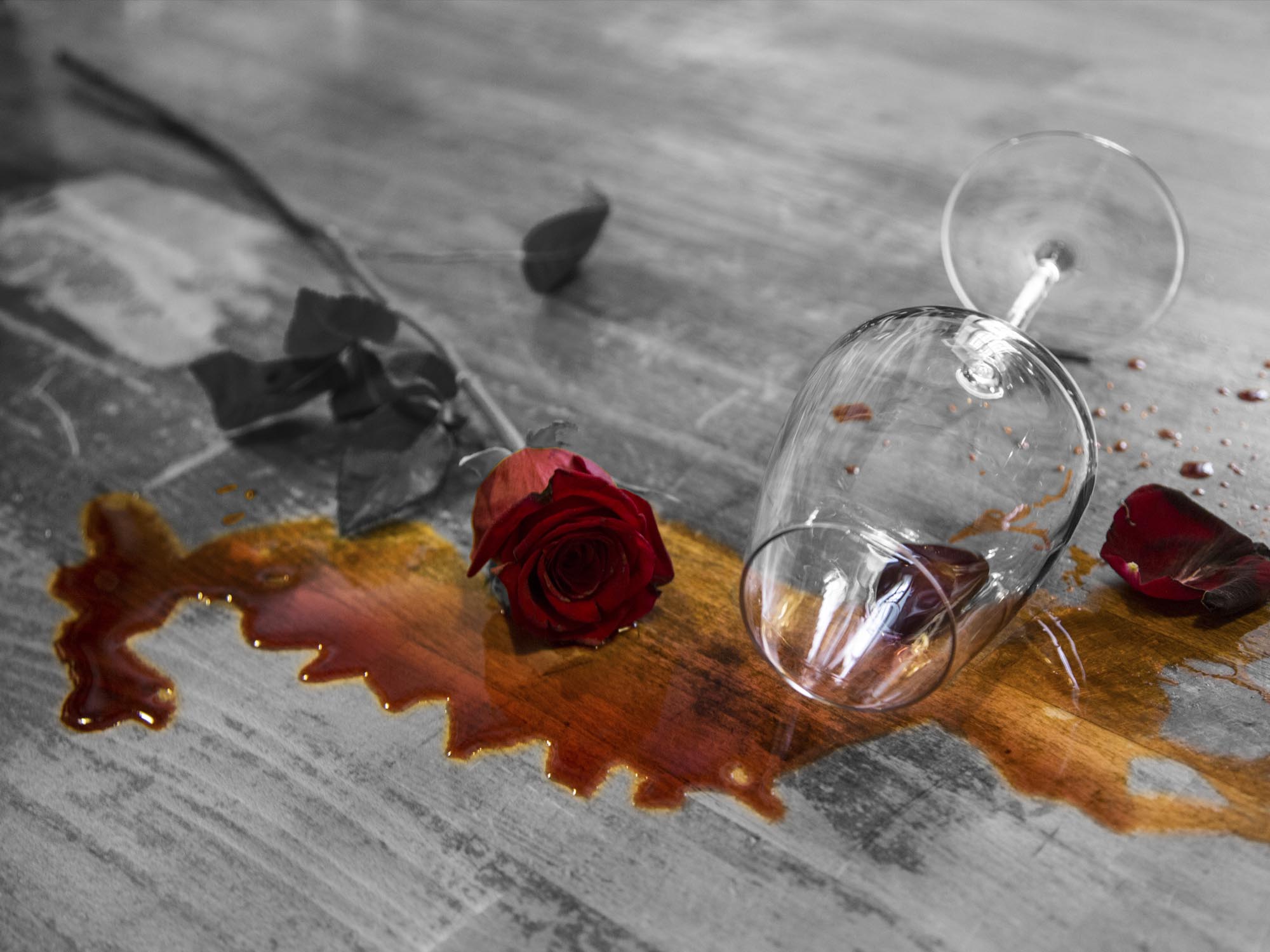 Still life in bianco e nero e rosso di un calice da vino ribaltato vicino a una rosa sopra dei parchetti di legno. Sotto c'è una macchia di vino.