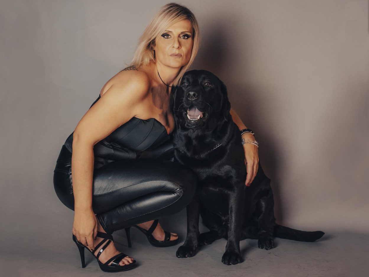 Una donna accucciata con un braccio attorno alla schiena di un cane nero di taglia grande